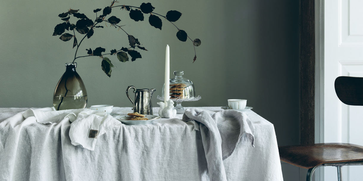 Dining table dressed in white distressed linen with plants and candles on top and a sage green backdrop