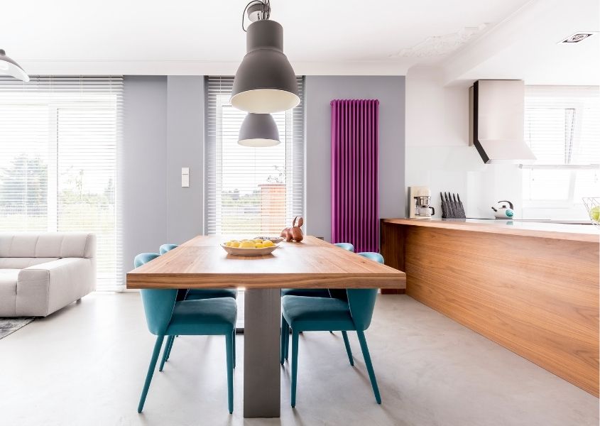 Open plan kitchen/diner with tall bright pink radiator and industrial dining table with green fabric dining chairs