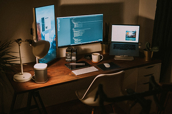 Computers on top of a dark wooden home office desk with a table lamp, a mug and a desk chair