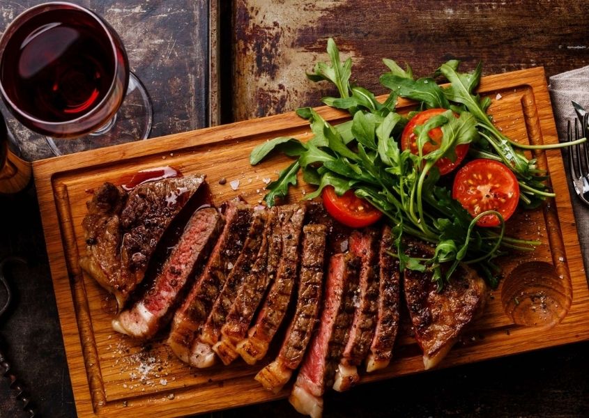 Slices of cooked steak on wooden board with green salad and glass of red wine
