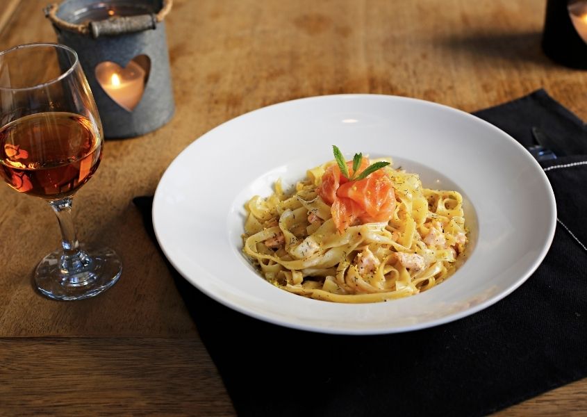 White bowl of salmon pasta on wooden dining table with black fabric plate mat and glass of wine