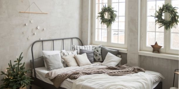 White bedroom with metal bedframe and hanging Christmas wreaths on window and small Christmas tree beside bed