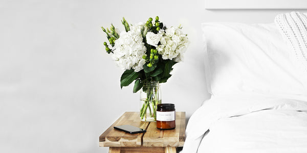 A white bouquet of flowers on a wooden bedside table next to a white bed
