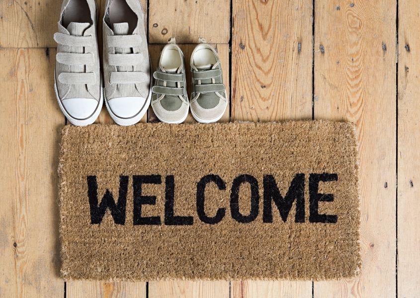 Hessian Welcome door mat with a grey adult pair of shoes and children's pair of shoes on wooden floor