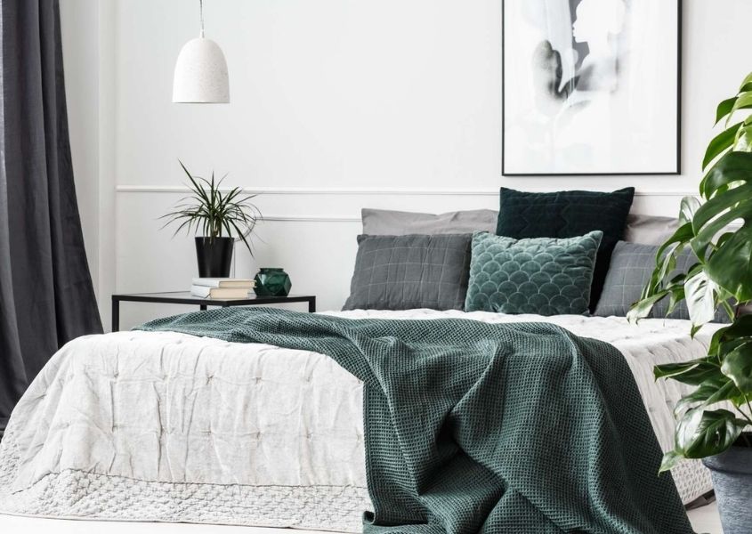 Large bed with grey blanket thrown over end and various grey coloured cushion. Bedroom also features a white hanging pendant light and large green house plant