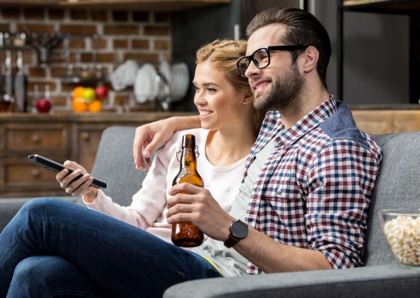 Couple sat on the sofa holding a bottle of beer and watching a film.