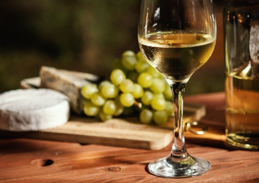 Glass of white wine on wooden table with selection of cheese and grapes in background