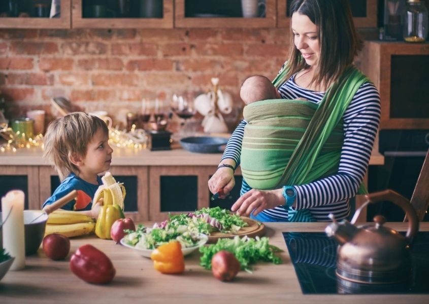 Women in rustic kitchen with infant in a baby carrier chopping vegetables with her toddler