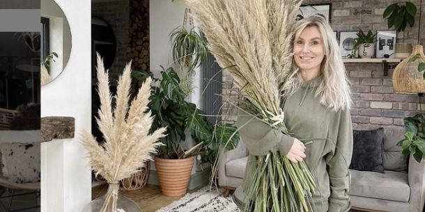 Woman with long blond hair and light green top carrying a large bunch of pampas grass