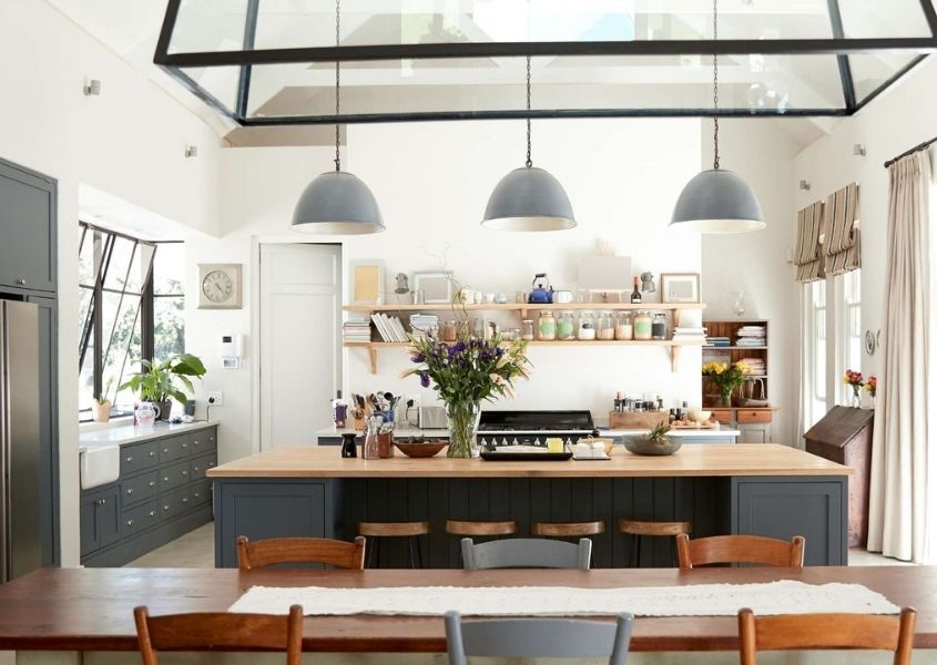 Large kitchen/diner with grey kitchen units, wooden dining table and three hanging ceiling lights in a room with a glassed pitched roof.