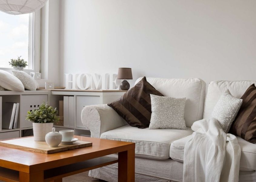 White living room with white fabric sofa, wooden coffee table and shelf with white painted wooden letters spelling Home