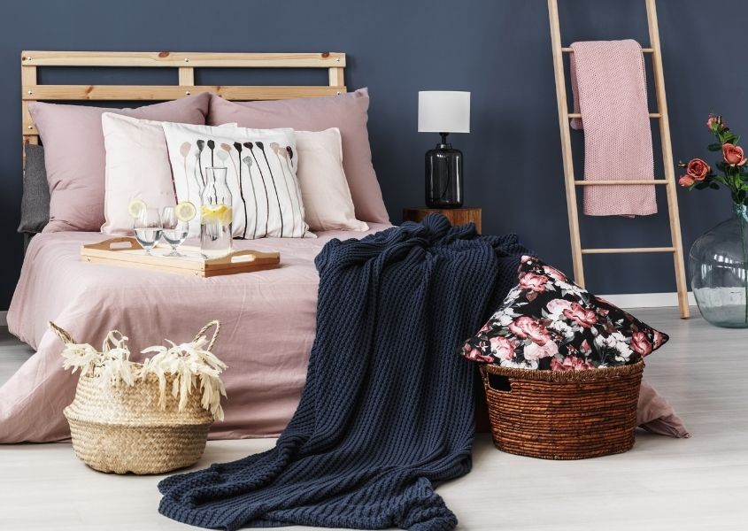 Bedroom with dark blue painted wall, wooden bed and wicker baskets on the floor with blue blanket hanging off floor.