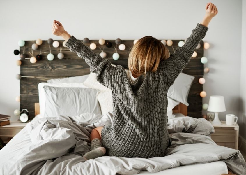 Woman in grey jumper stretching on wooden double bed with fairy lights on headboard