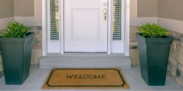 Welcome doormat in front of white front door with two large grey plant pots with green shrubs either side
