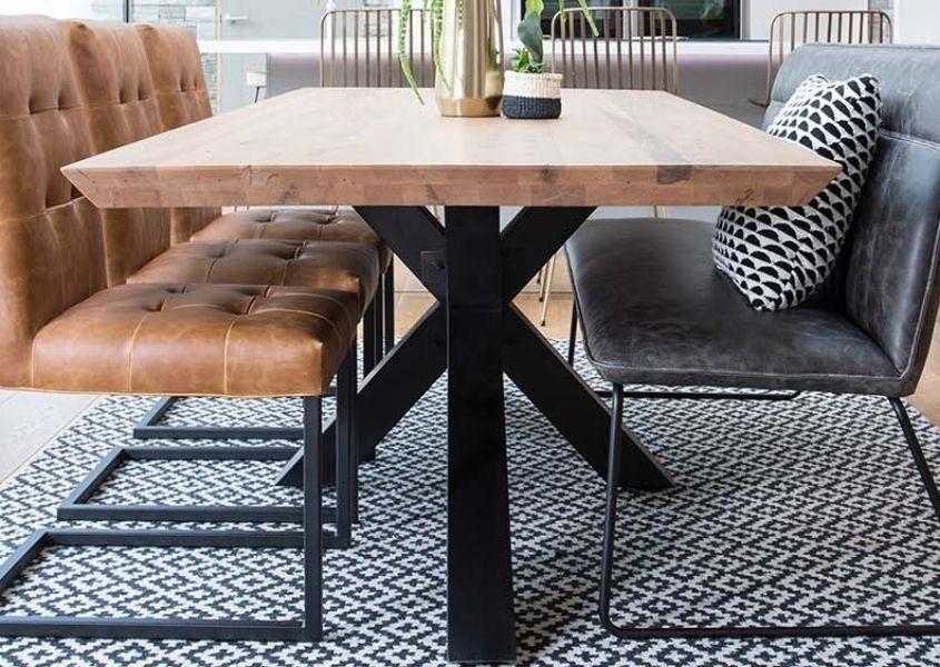 Close up of wooden dining table with thick black metal spider leg, tan leather dining chairs and grey faux leather bench with back