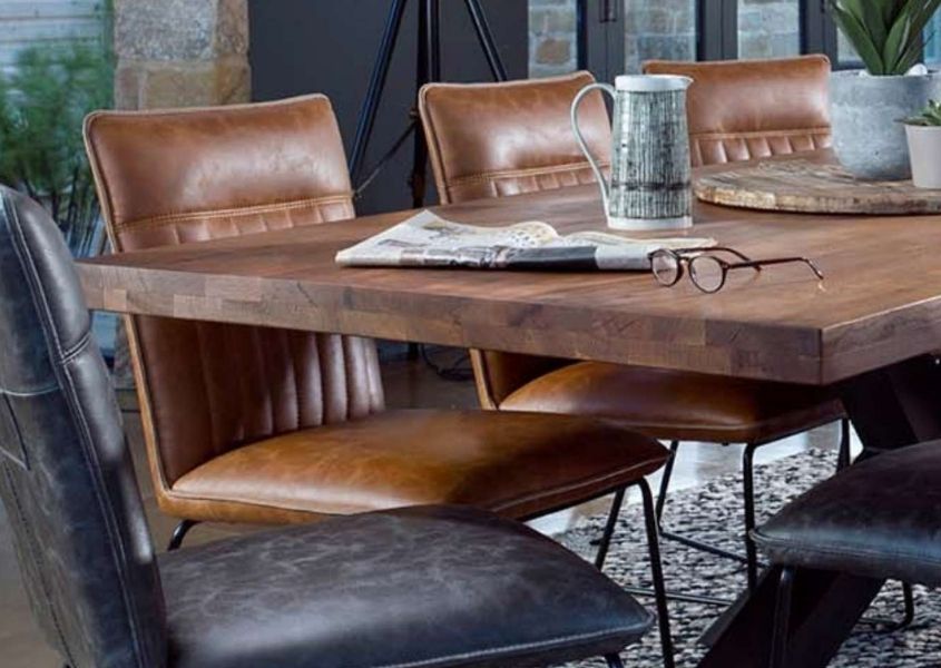 Corner of a rustic dining table with a brown and grey faux leather dining chairs and newspaper and spectacles on table top