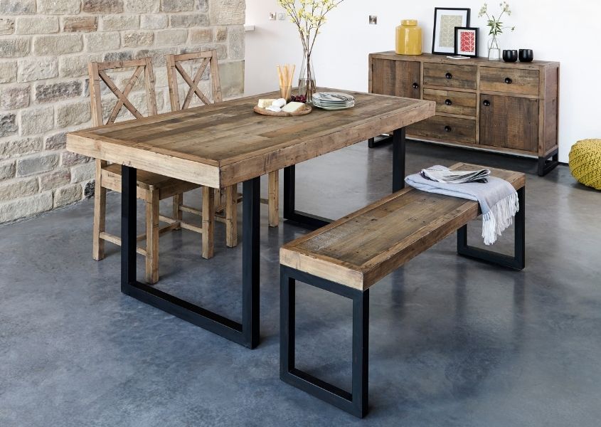 Industrial dining table in reclaimed wood with black u-shaped frame and matching wooden bench. Also features two wooden dining chairs and set in a room with an exposed brick wall
