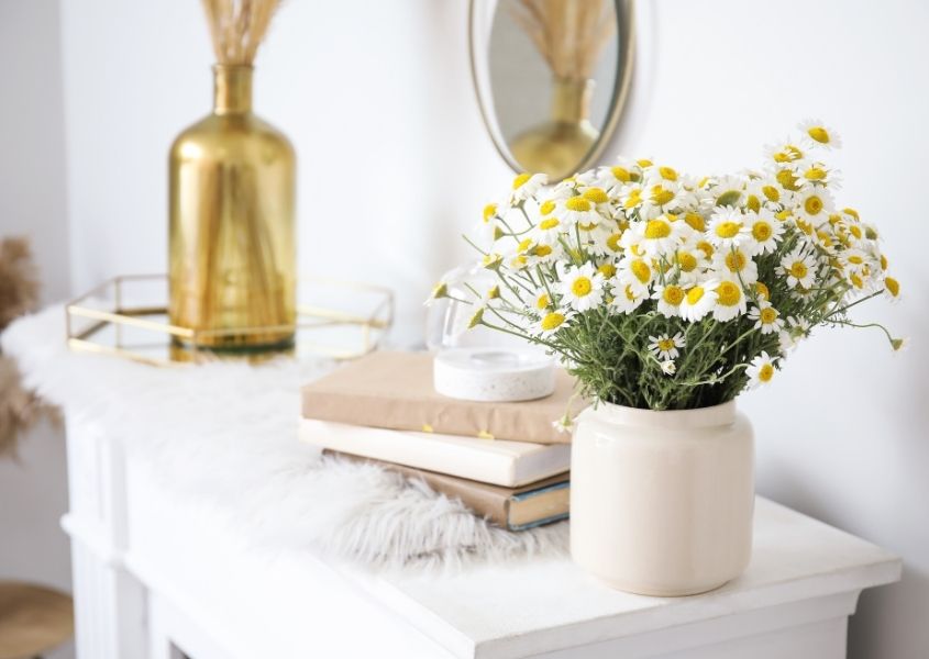 White and yellow flowers in white vase on a fireplace with gold vase and books