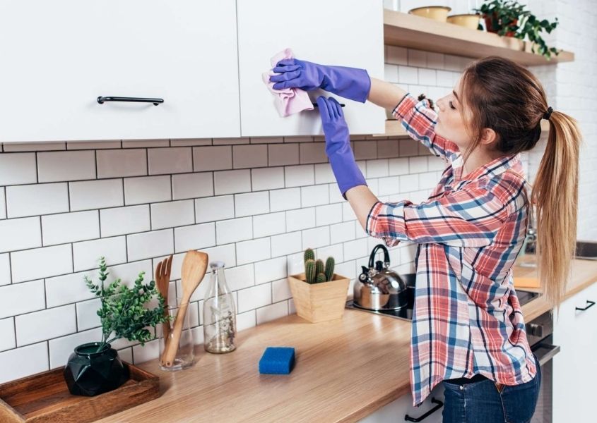 Woman wearing a checked shirt and purple rubber gloves cleaning white kitchen units