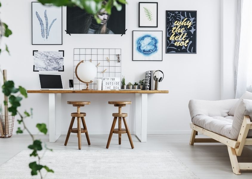 Home office desk with two wooden stools and white sofa with green plants on art on wall
