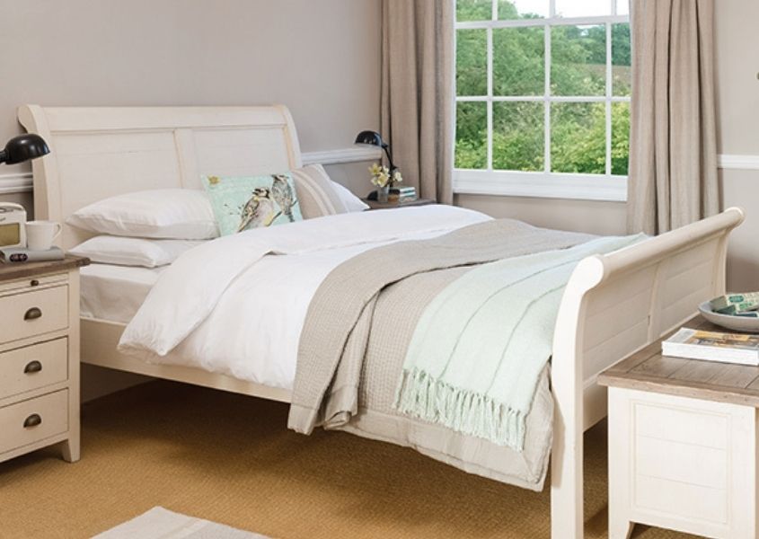 White wooden king size bed with white covers, white painted bedside table and blanket box and beige curtains
