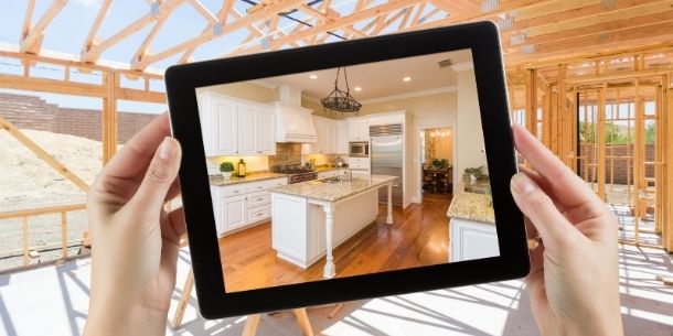 Hands holding a tablet screen with photo of a white kitchen against a background of a building site with wooden beams and frame
