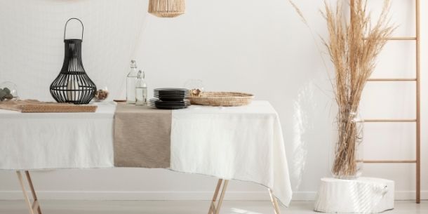White dining room with wooden table and white and beige table cloth and tall display of dried palms in corner of room
