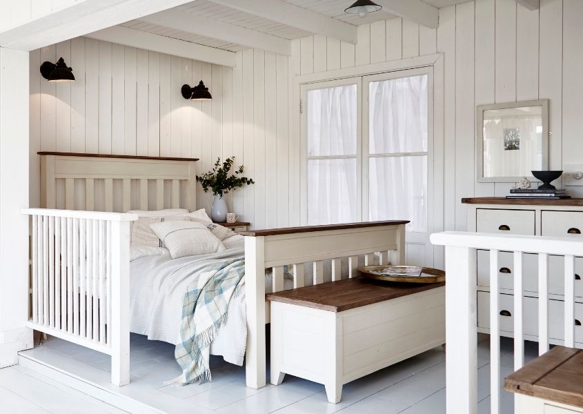 White painted reclaimed wood bed in white wood panelled bedroom with matching wooden blanket box and black spot lights on wall behind bed